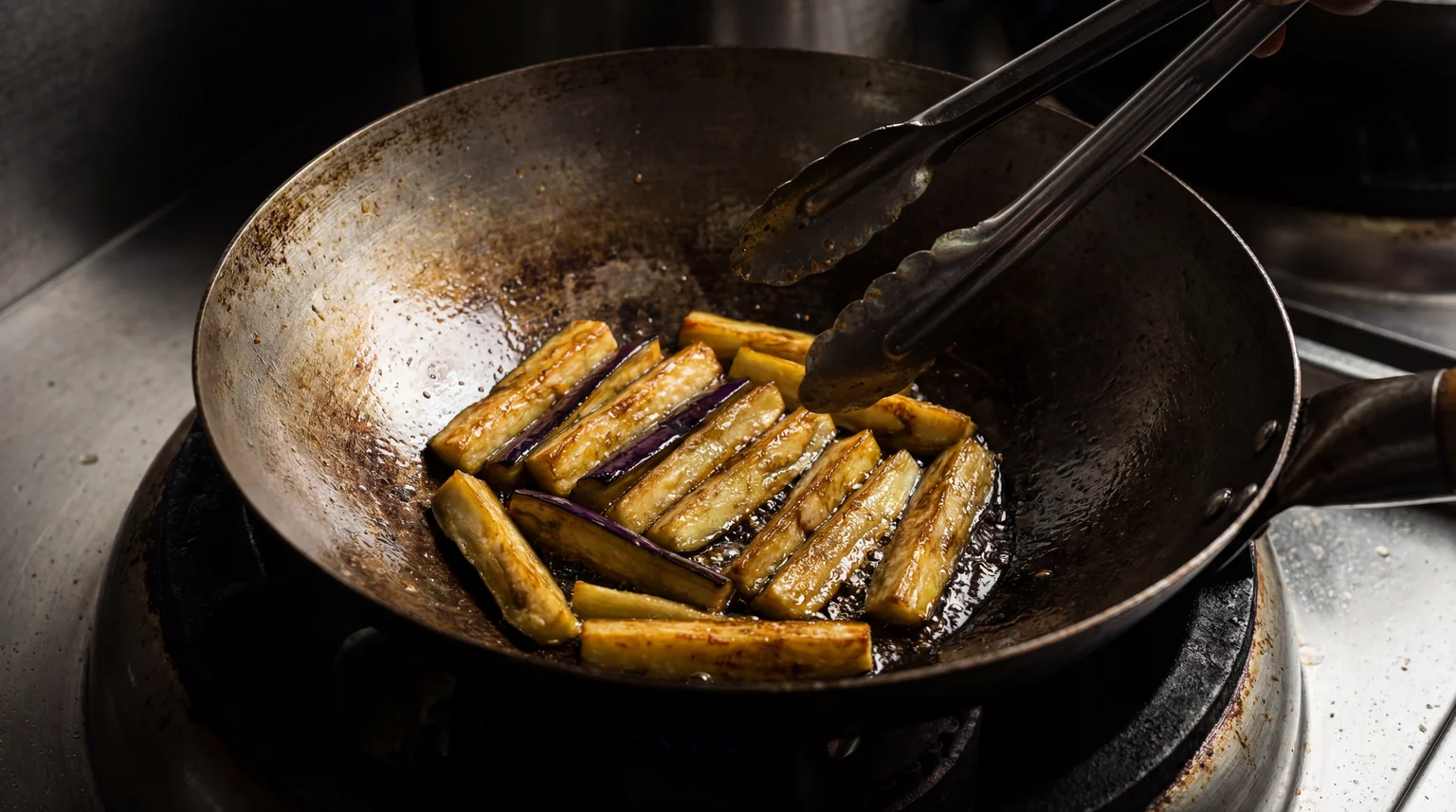 Golden-brown Chinese eggplant batons sizzling in a seasoned carbon steel wok, held with metal tongs
