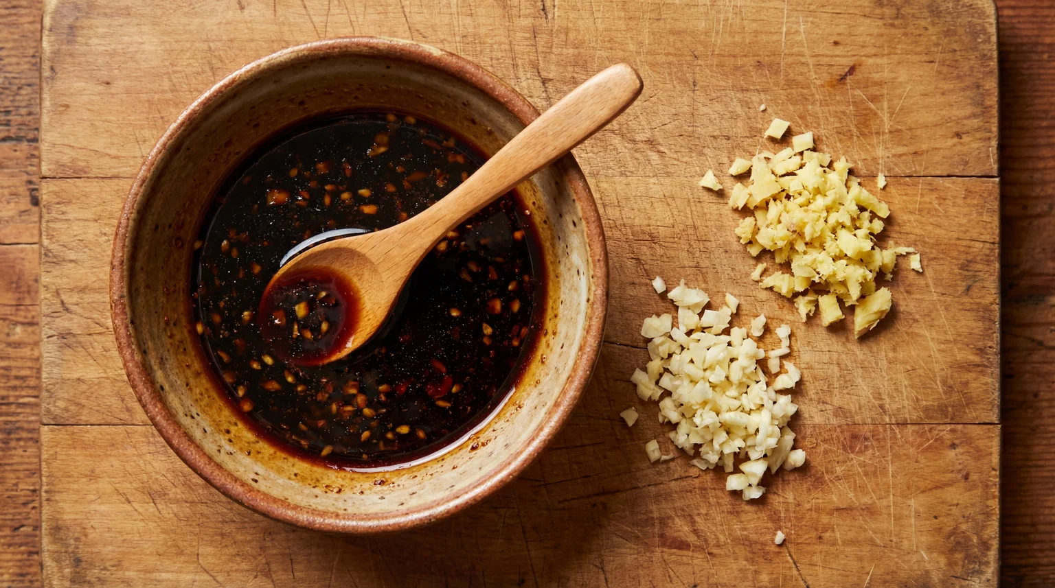 Dark garlic sauce in a ceramic bowl with wooden spoon, minced garlic and ginger on a wooden cutting board