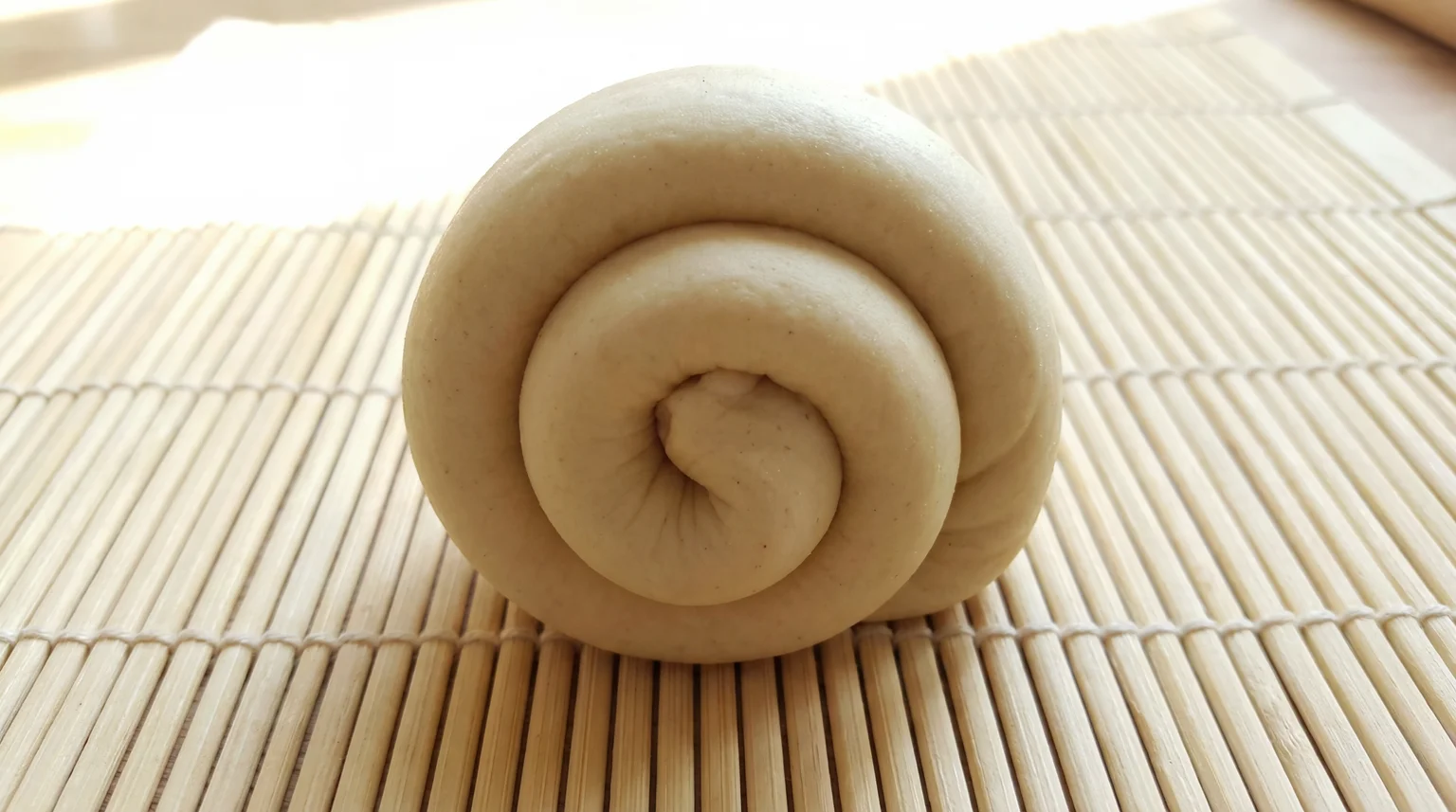 Coiled scallion pancake dough in spiral snail shape resting on bamboo mat, traditional lamination technique for flaky layers