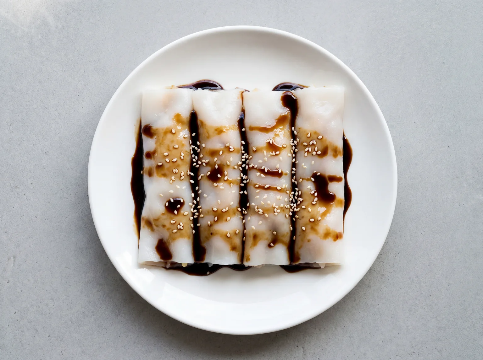 Four freshly rolled cheung fun rice noodle rolls on a white plate drizzled with dark soy sauce and sesame seeds