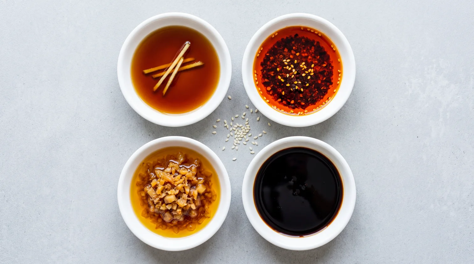 Four dim sum dipping sauces in white ceramic bowls including soy ginger, chilli oil, XO sauce and sweet soy on grey concrete