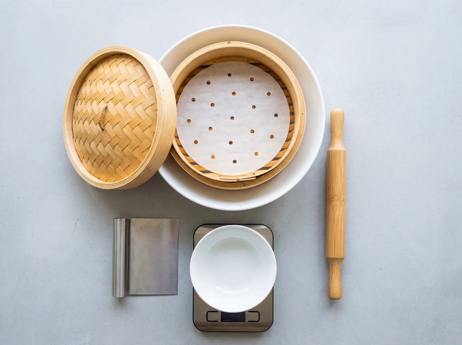 Bamboo steamer basket, rolling pin, kitchen scale and pastry scraper laid out on grey concrete for making dim sum at home
