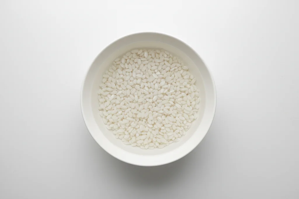 Overhead close-up of white glutinous rice grains soaking in clear still water in a white ceramic bowl