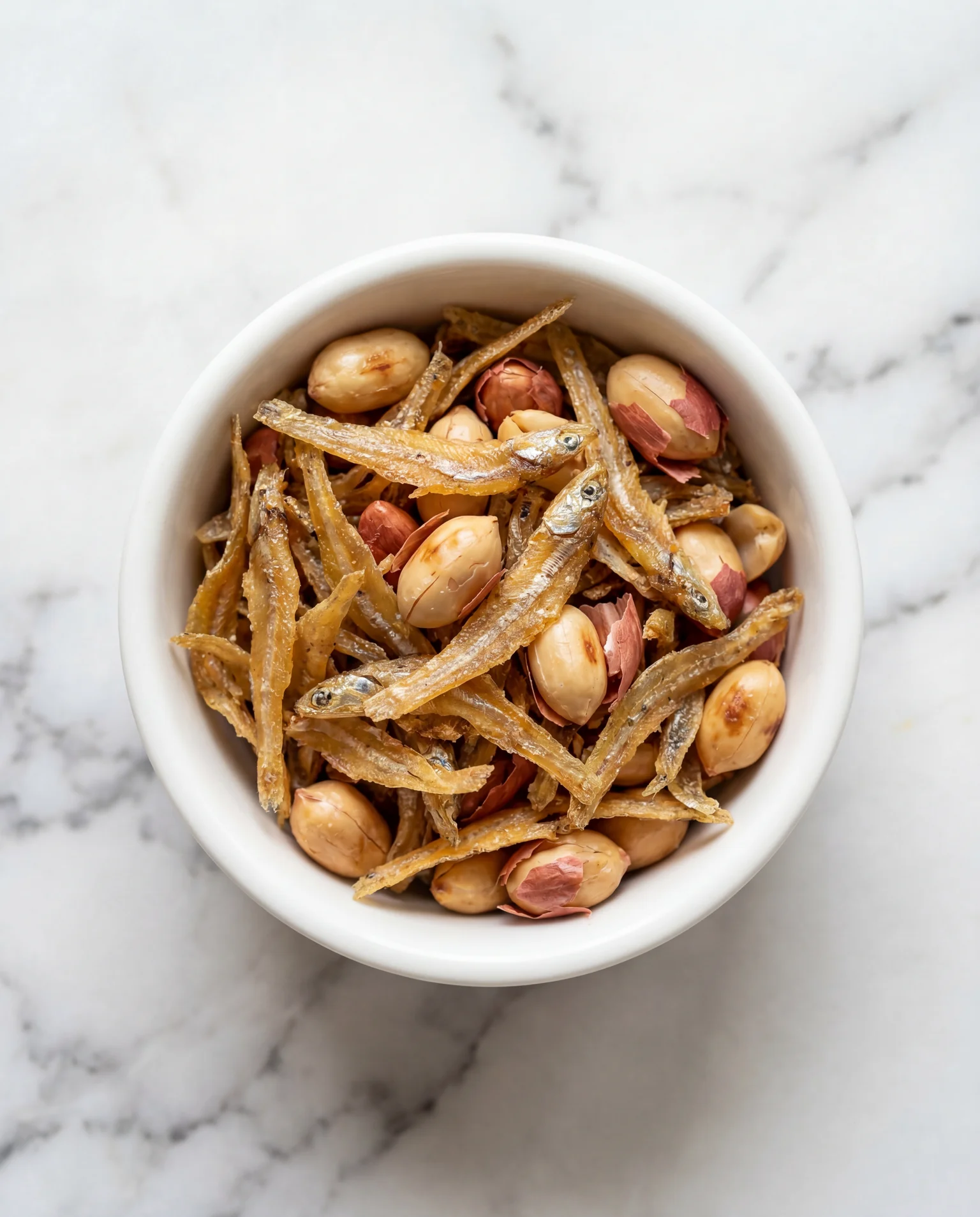 Golden crispy ikan bilis (fried dried anchovies) and roasted peanuts in a white ceramic bowl