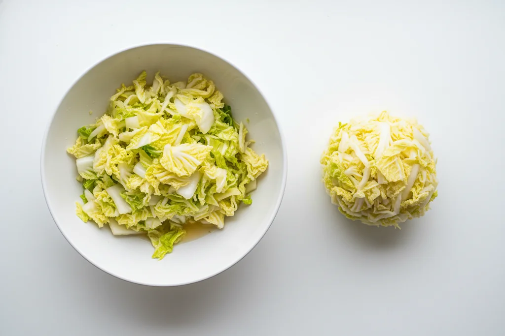 Salted napa cabbage in white bowl beside hand-squeezed dry cabbage ball on seamless white surface