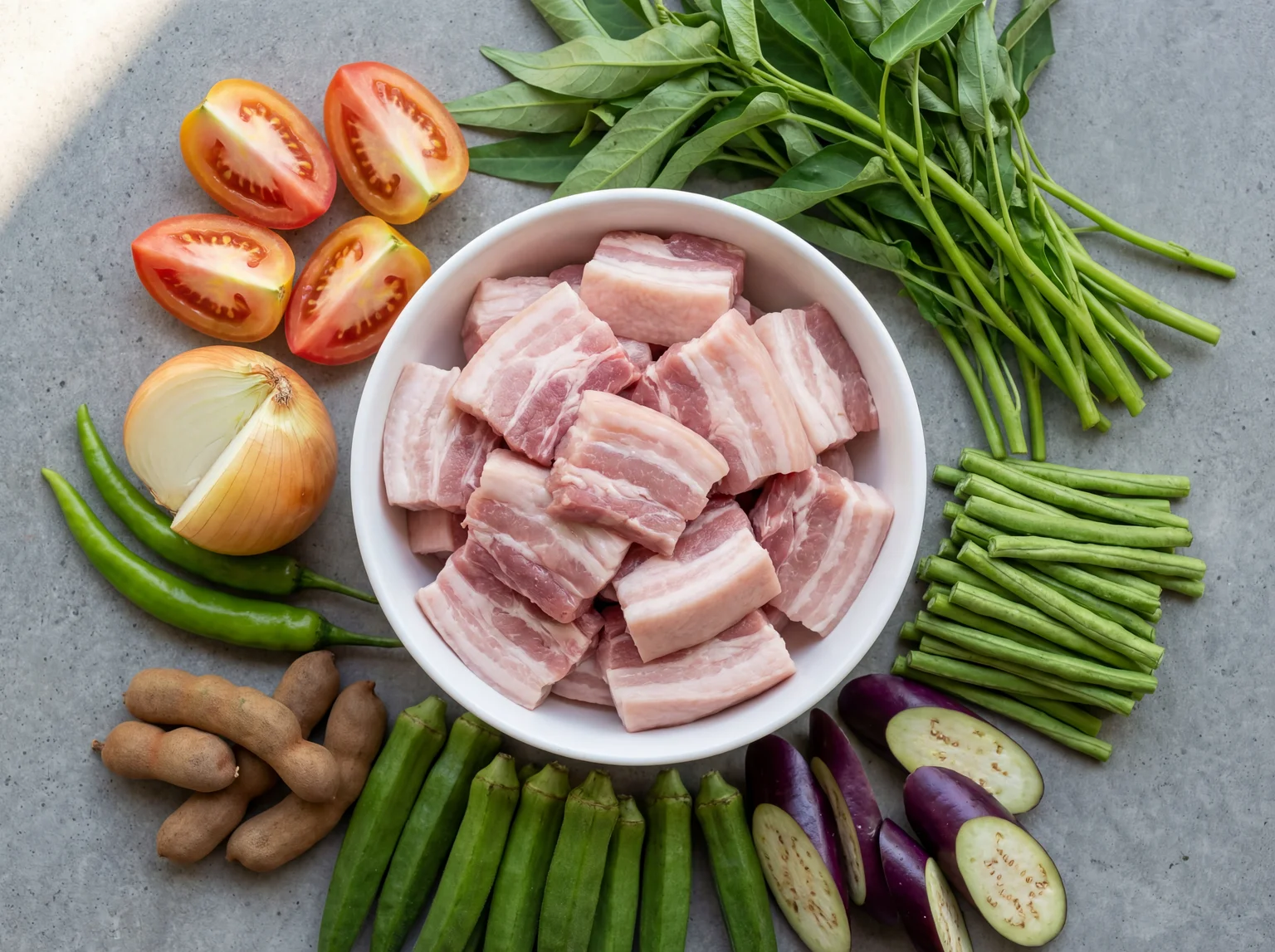 Raw ingredients for Sinigang na Baboy — pork belly chunks, tamarind pods, kangkong, eggplant, okra, sitaw, tomatoes, and onion staged in a white ceramic bowl