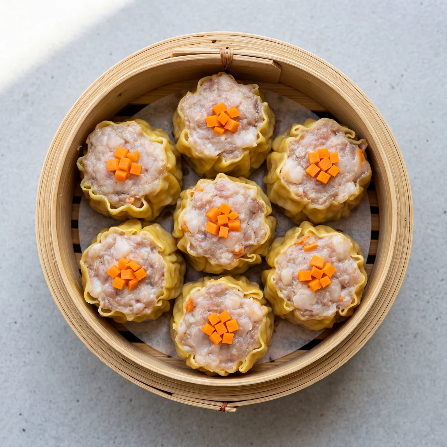Six open-topped siu mai dumplings with yellow wonton wrappers and pork prawn filling topped with orange carrot in a bamboo steamer