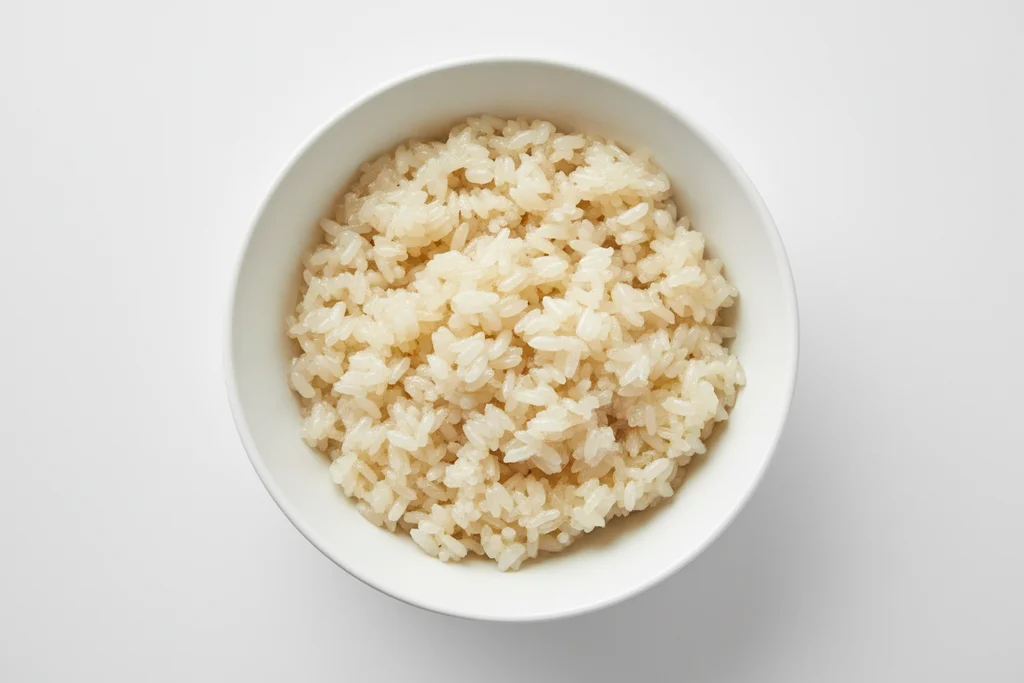 Overhead close-up of glossy coconut-soaked sticky rice grains in a white bowl, plump and ivory-coloured after absorbing the coconut sauce