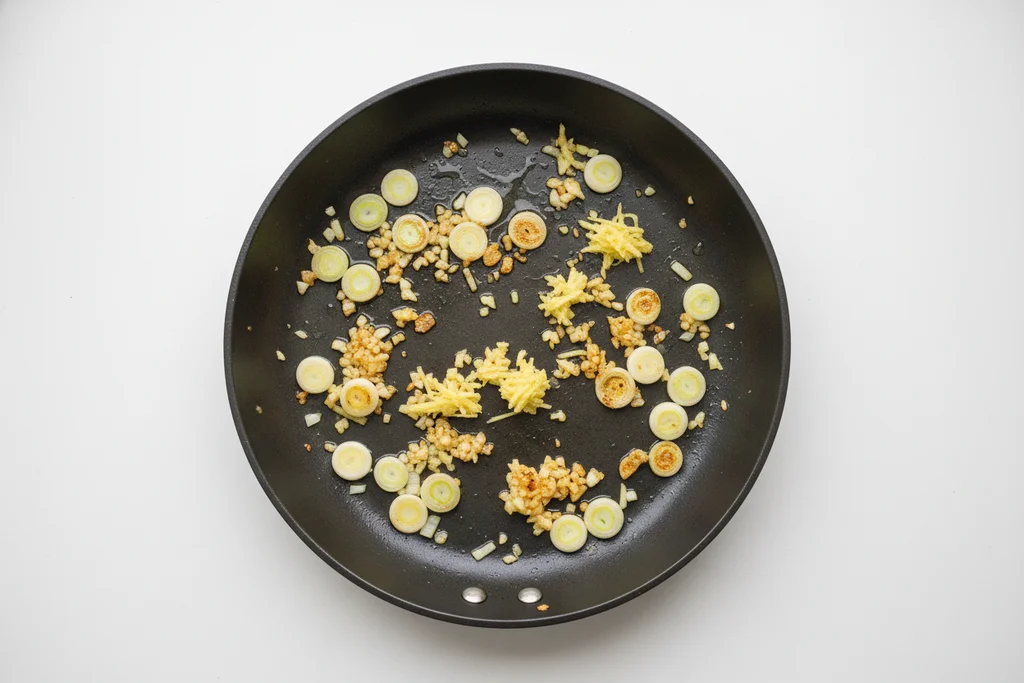 Minced garlic turning golden, grated ginger and sliced green onion whites sizzling in a hot dark skillet — the aromatic base for shrimp and bell pepper stir fry